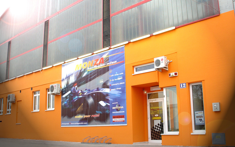 Leuchtend orangefarbenes Gebäude mit mehreren Fenstern, einer Glastür und einem großen Werbeschild der Monza Kartbahn Wien, auf dem ein fahrender Rennwagen abgebildet ist.