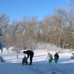 Mehrere Personen befinden sich im Freien in einem verschneiten Park; ein Erwachsener hilft einem Kind auf einen Schlitten, während zwei weitere Personen bei Schneetreiben im Hintergrund bergauf gehen.