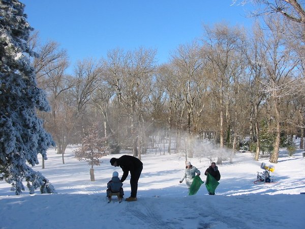 Mehrere Personen befinden sich im Freien in einem verschneiten Park; ein Erwachsener hilft einem Kind auf einen Schlitten, während zwei weitere Personen bei Schneetreiben im Hintergrund bergauf gehen.