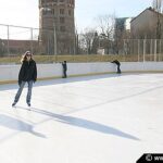 Mehrere Personen, die bei Tageslicht auf einer Freiluft-Eisbahn Schlittschuh laufen, mit Gebäuden und Bäumen im Hintergrund.