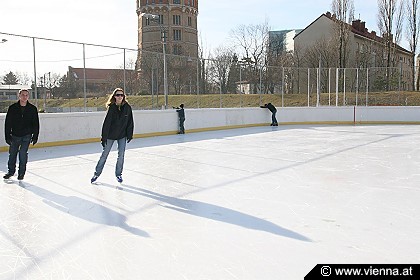Mehrere Personen, die bei Tageslicht auf einer Freiluft-Eisbahn Schlittschuh laufen, mit Gebäuden und Bäumen im Hintergrund.