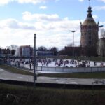 Menschen laufen Schlittschuh auf einer Eisbahn im Freien in der Nähe eines hohen historischen Turms mit Kuppeldach unter einem teilweise bewölkten Himmel.