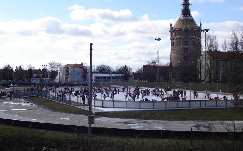 Menschen laufen Schlittschuh auf einer Eisbahn im Freien in der Nähe eines hohen historischen Turms mit Kuppeldach unter einem teilweise bewölkten Himmel.