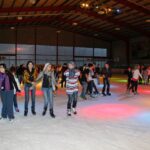 Eine Gruppe von Menschen, die in einer Eishalle unter bunten Lichtern Schlittschuh laufen; einige halten sich an den Händen, während andere einzeln laufen.