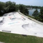 Ein Beton-Skatepark mit mehreren Rampen und Bowls befindet sich an einem See, umgeben von Gras, Bäumen und einem Spazierweg.