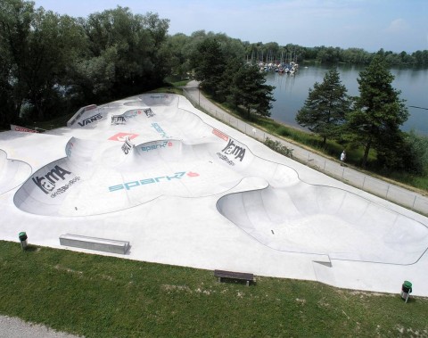 Ein Beton-Skatepark mit mehreren Rampen und Bowls befindet sich an einem See, umgeben von Gras, Bäumen und einem Spazierweg.