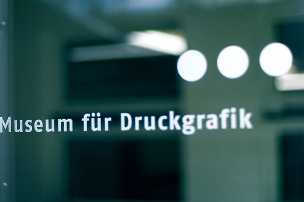 Glastür mit der Aufschrift "Museum für Druckgrafik" in deutscher Sprache, mit drei weißen Punkten über dem Wort "Museum" und einem unscharfen inneren Hintergrund.