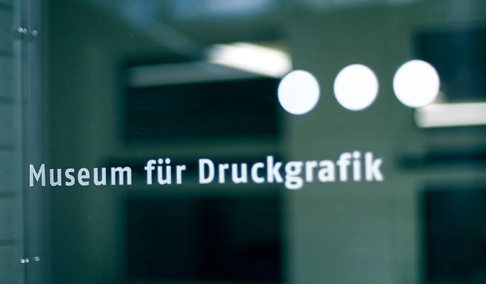 Glastür mit der Aufschrift "Museum für Druckgrafik" in deutscher Sprache, mit drei weißen Punkten über dem Wort "Museum" und einem unscharfen inneren Hintergrund.