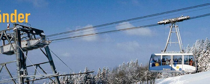 Die Seilbahn mit der Aufschrift "Pfänder" fährt über verschneite Bäume und Landschaften unter einem klaren blauen Himmel; die Einstiegsstation ist links zu sehen.