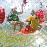 Zwei Personen befinden sich in einem großen, transparenten, aufblasbaren Zorb-Ball mit roten Gurten und rollen auf einer Wiese unter blauem Himmel.