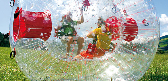 Zwei Personen befinden sich in einem großen, transparenten, aufblasbaren Zorb-Ball mit roten Gurten und rollen auf einer Wiese unter blauem Himmel.