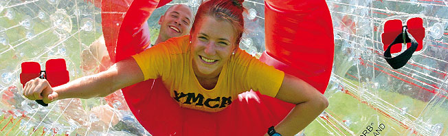 Zwei Personen lächeln, während sie sich in einem durchsichtigen, aufblasbaren Zorb-Ball befinden, wobei die vordere Person ein gelbes YMCA-Shirt trägt und rote Griffe hält.
