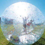 Zwei Personen in einem großen, durchsichtigen, aufblasbaren Ball, einem so genannten Zorb, rollen auf einer Wiese unter einem strahlend blauen Himmel.