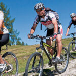 Fünf Radfahrer fahren an einem sonnigen Tag mit Bäumen und blauem Himmel im Hintergrund auf einem grasbewachsenen, felsigen Weg Mountainbike.