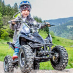 Ein kleines Kind mit Helm fährt mit einem kleinen vierrädrigen ATV auf einem unbefestigten Weg in einem grasbewachsenen, bergigen Außenbereich.