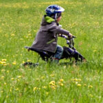 Ein Kind, das einen Helm trägt, fährt mit einem kleinen Fahrrad durch eine grasbewachsene Wiese mit gelben Blumen.