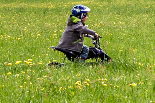 Ein Kind, das einen Helm trägt, fährt mit einem kleinen Fahrrad durch eine grasbewachsene Wiese mit gelben Blumen.