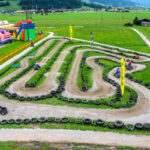 Eine kurvenreiche Kartbahn mit mehreren Personen, die Karts fahren, umrandet von Reifen, auf einer Wiese mit Spielgeräten und Bergen im Hintergrund.