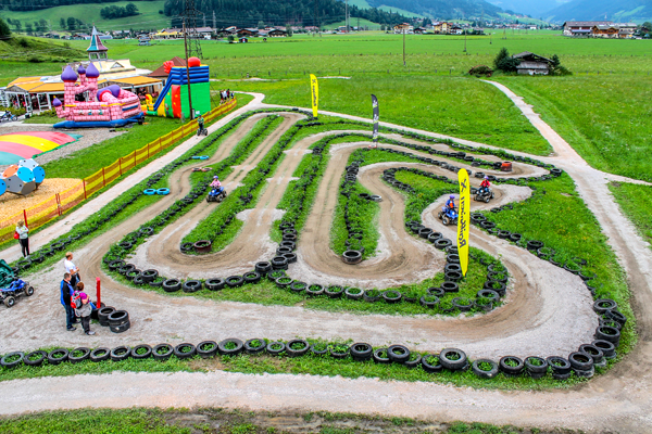 Eine kurvenreiche Kartbahn mit mehreren Personen, die Karts fahren, umrandet von Reifen, auf einer Wiese mit Spielgeräten und Bergen im Hintergrund.
