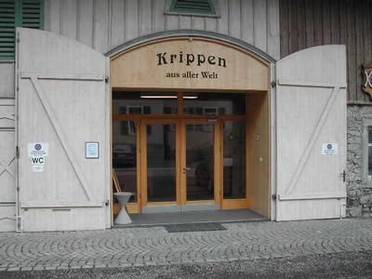 Eingang zu einem Gebäude mit großen Holztüren, Glaseingang und einem Schild darüber, auf dem „Krippen aus aller Welt“ steht. An der Fassade sind Hinweisschilder für WC und Museum zu sehen.