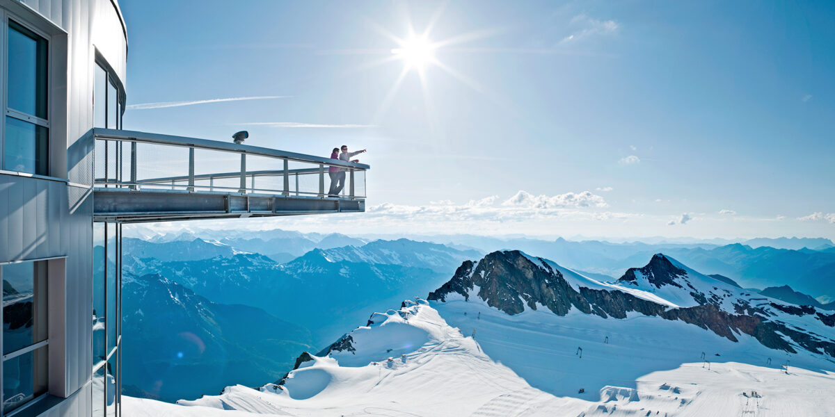 Eine Aussichtsplattform erstreckt sich von einem modernen Gebäude aus über verschneite Berge bei strahlendem Sonnenschein, und zwei Personen betrachten die Landschaft.