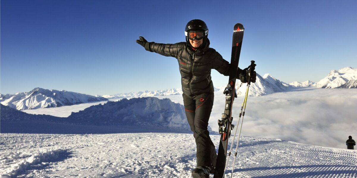 Eine Person in Skikleidung steht auf einem verschneiten Berghang, hält Skier und lächelt mit ausgestreckten Armen, mit schneebedeckten Bergen und einem klaren Himmel im Hintergrund.