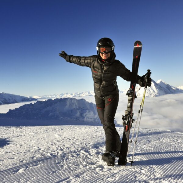Eine Person in Skikleidung steht auf einem verschneiten Berghang, hält Skier und lächelt mit ausgestreckten Armen, mit schneebedeckten Bergen und einem klaren Himmel im Hintergrund.