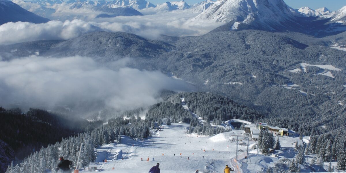 Skifahrer fahren einen verschneiten Berghang hinunter, der von Kiefern umgeben ist. Im Hintergrund sind ein Sessellift und die Alpengipfel bei klarem Himmel zu sehen.