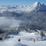 Skifahrer fahren einen verschneiten Berghang hinunter, der von Kiefern umgeben ist. Im Hintergrund sind ein Sessellift und die Alpengipfel bei klarem Himmel zu sehen.