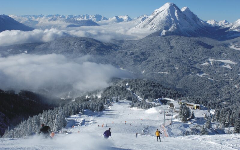 Skifahrer fahren einen verschneiten Berghang hinunter, der von Kiefern umgeben ist. Im Hintergrund sind ein Sessellift und die Alpengipfel bei klarem Himmel zu sehen.