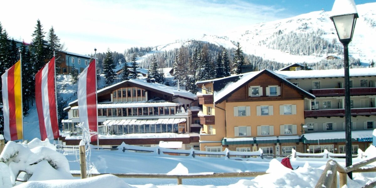 Schneebedeckte Gebäude und Kiefern stehen vor einem Berghang unter klarem Himmel; im Vordergrund sind mehrere senkrechte rote, gelbe und weiße Fahnen zu sehen.