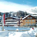 Schneebedeckte Gebäude und Kiefern stehen vor einem Berghang unter klarem Himmel; im Vordergrund sind mehrere senkrechte rote, gelbe und weiße Fahnen zu sehen.