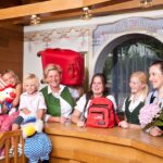 Fünf Frauen und zwei Kinder stehen lächelnd hinter einem hölzernen Empfangstresen. Die Kinder halten Plüschtiere in der Hand. Gepäck und ein Blumenstrauß sind zu sehen. Die Umgebung wirkt einladend und fröhlich.