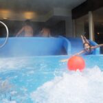 Ein Kind rutscht auf einer blauen Wasserrutsche in ein Becken und greift nach einem roten Ball im Wasser, während andere Kinder oben auf der Rutsche stehen.
