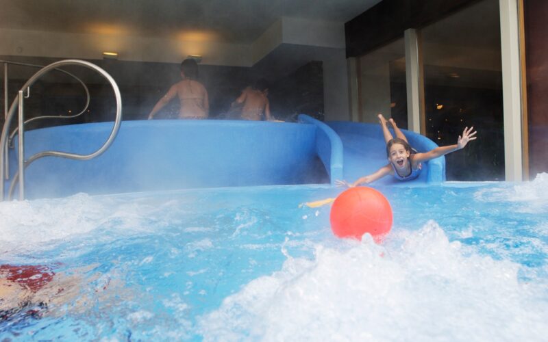 Ein Kind rutscht auf einer blauen Wasserrutsche in ein Becken und greift nach einem roten Ball im Wasser, während andere Kinder oben auf der Rutsche stehen.