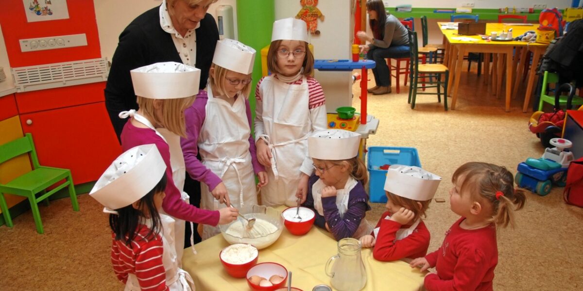 Eine Gruppe junger Kinder mit Schürzen und Kochmützen mischt in einem farbenfrohen Klassenzimmer unter der Aufsicht eines Erwachsenen Zutaten in Schüsseln an einem Tisch.