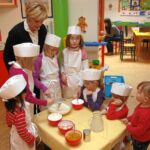 Eine Gruppe junger Kinder mit Schürzen und Kochmützen mischt in einem farbenfrohen Klassenzimmer unter der Aufsicht eines Erwachsenen Zutaten in Schüsseln an einem Tisch.