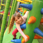 Ein junges Mädchen lächelt, während es auf ein großes, grünes, mit Stacheln versehenes Hindernis in einem von Netzen umgebenen Indoor-Spielbereich klettert.