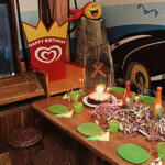 Piratengeburtstagsfeier mit dekoriertem Tisch, Kuchen mit Kerzen, grünen Tellern und Tassen, Luftschlangen und Piratendekoration im Hintergrund.