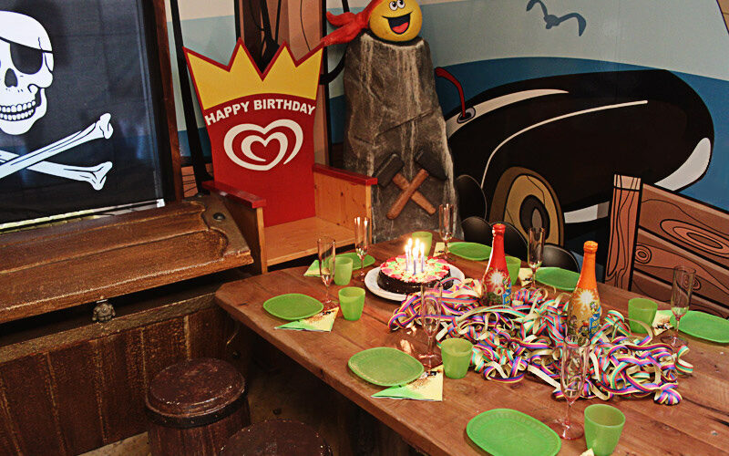 Piratengeburtstagsfeier mit dekoriertem Tisch, Kuchen mit Kerzen, grünen Tellern und Tassen, Luftschlangen und Piratendekoration im Hintergrund.