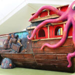 Ein farbenfroher Indoor-Spielplatz in Form eines hölzernen Piratenschiffs mit Fässern und einem großen lila Oktopus auf der Spitze.