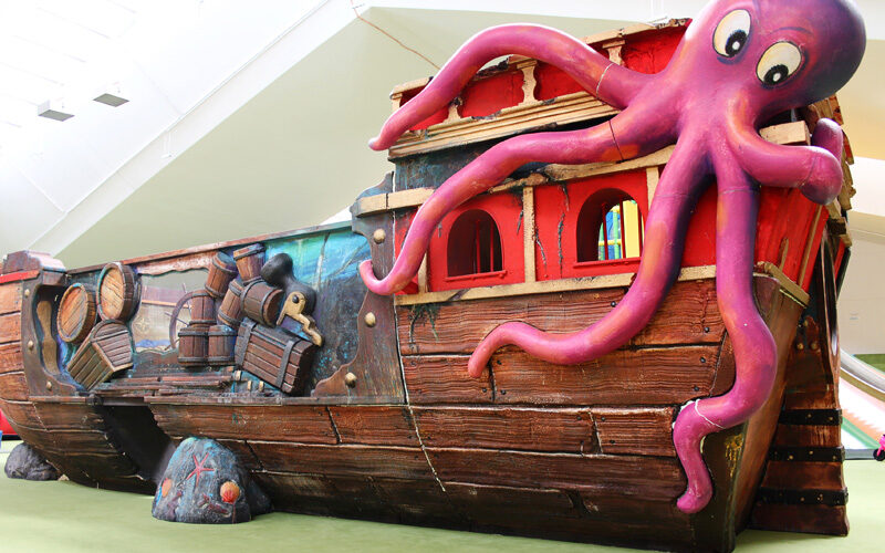 Ein farbenfroher Indoor-Spielplatz in Form eines hölzernen Piratenschiffs mit Fässern und einem großen lila Oktopus auf der Spitze.