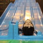 Ein Kind mit geflochtenen Haaren und schwarzem Hemd lächelt, während es auf dem Boden einer blauen Rutsche in einem Indoor-Spielplatz liegt.