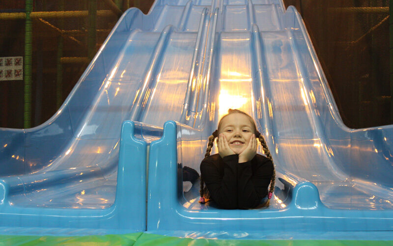 Ein Kind mit geflochtenen Haaren und schwarzem Hemd lächelt, während es auf dem Boden einer blauen Rutsche in einem Indoor-Spielplatz liegt.