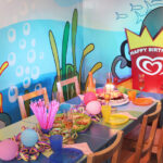 Ein dekorierter Geburtstagstisch mit Kuchen, Luftballons, Partyhüten, Getränken, Snacks und einem "Happy Birthday"-Schild vor einer farbenfrohen Wandmalerei mit Meeresmotiven.