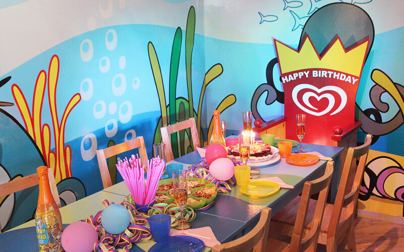 Ein dekorierter Geburtstagstisch mit Kuchen, Luftballons, Partyhüten, Getränken, Snacks und einem "Happy Birthday"-Schild vor einer farbenfrohen Wandmalerei mit Meeresmotiven.