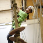 Ein Kind, das einen Helm und einen Klettergurt trägt, lächelt beim Klettern auf einem Seilhindernisparcours in einer Halle.