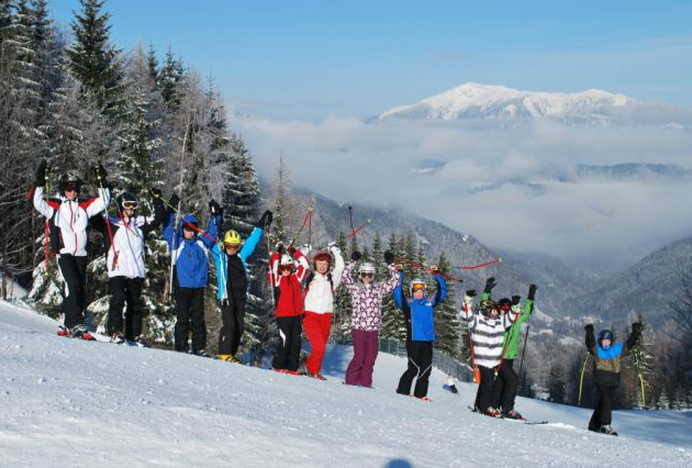 Eine Gruppe von Menschen in Winterkleidung steht mit erhobenen Skistöcken auf einem verschneiten Hang, umgeben von Bäumen und Bergen unter einem klaren Himmel.