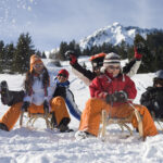 Sechs Personen in Winterkleidung fahren auf Holzschlitten einen verschneiten Hügel hinunter, im Hintergrund sind Bäume und ein schneebedeckter Berg zu sehen.