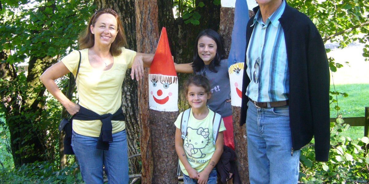 Vier Personen stehen im Freien in der Nähe von Bäumen und drei hohen Holzstatuen, die als lächelnde Gnome mit roten und blauen Hüten bemalt sind.